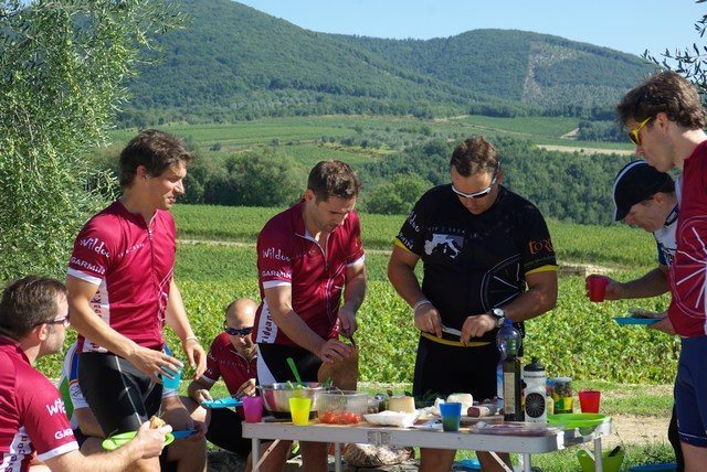 Seven people dressed in cyclist clothing gather around a picnic table in a vineyard, laid with picnic food.