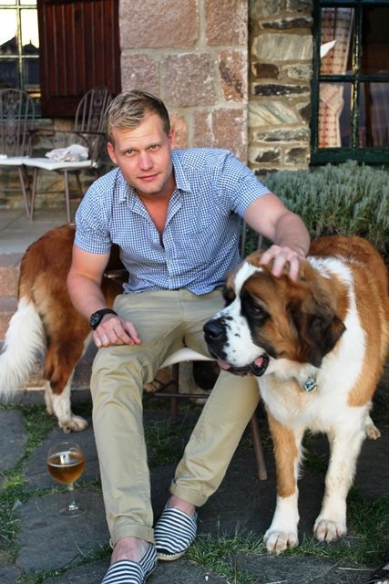 A man is seated on a patio, patting a large brown and white dog. The tail end of another dog can be seen behind him.