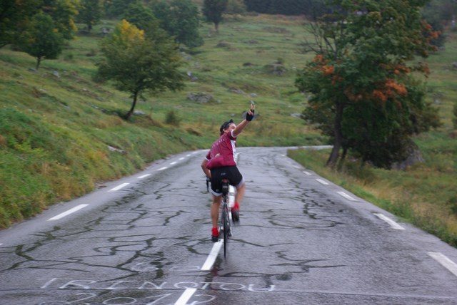 A cyclist rides away, down a paved road waving back to the camera.