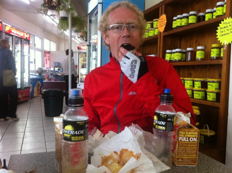 A man in a red cycling coat eating. There is more food in front of him on a table.