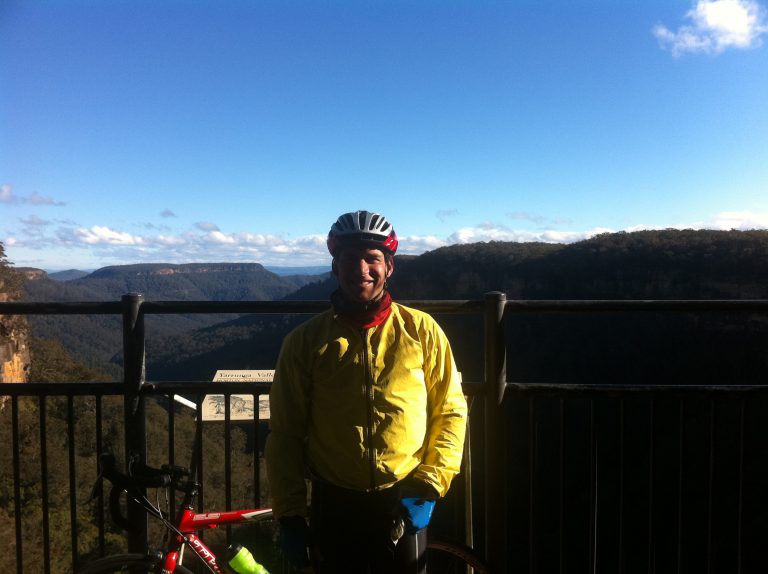 A cyclist wearing bright warm cycling clothes stands posing in front of a sweeping view of a valley.