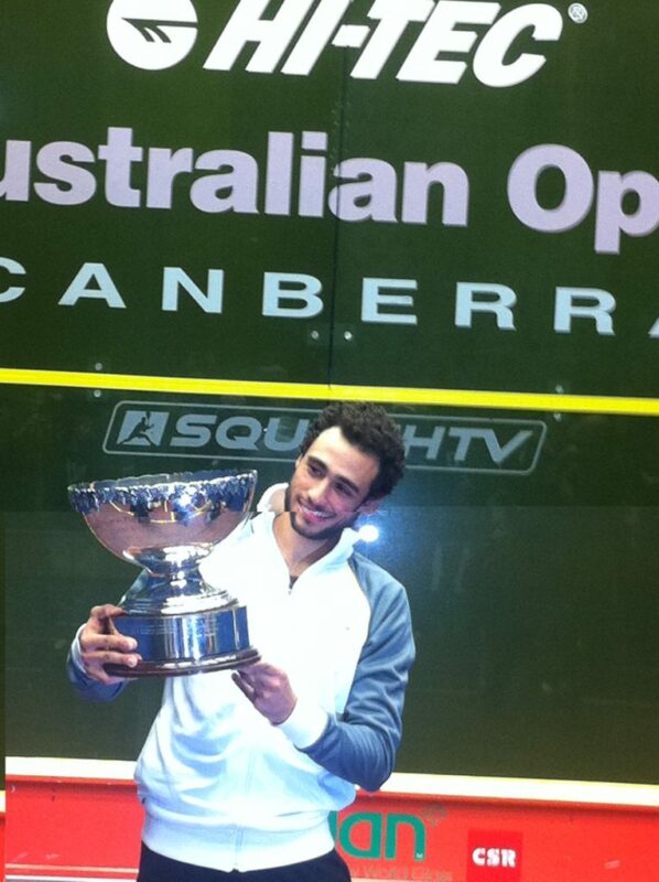 A man in tennis clothing holds a large, silver trophy, standing in front of a huge banner.