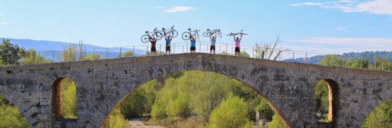 Cyclists on Ride and Seek bike tours holding their bikes above their heads, standing on an old Roman stone bridge in Provence.