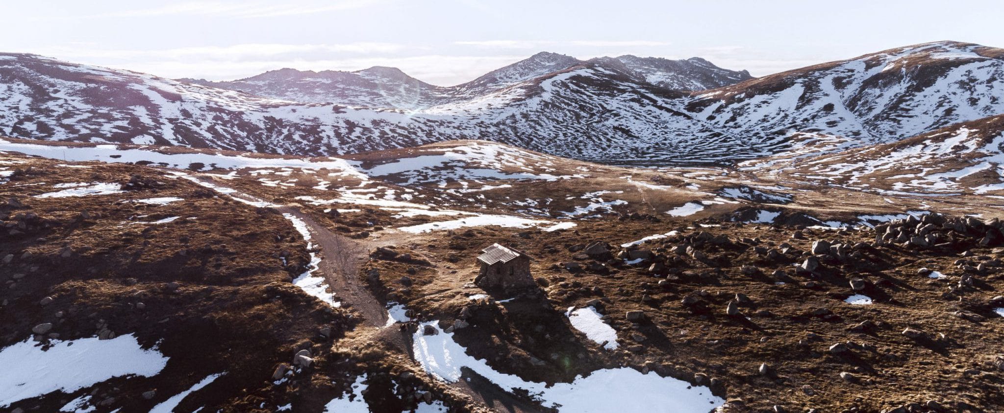 Looking across the high plains over some small lakes in the foreground and snow-covered peaks in the background. A sole highcountry hut sits in the centre beside one of the lakes.