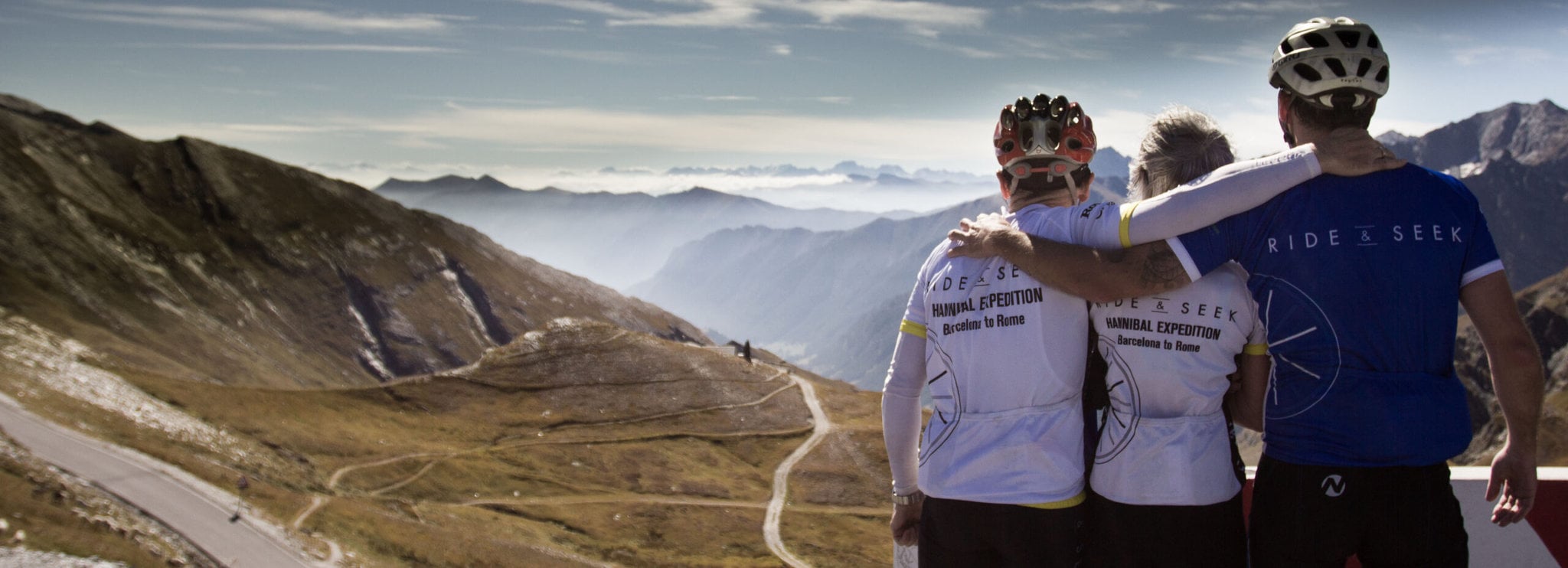 Three people stand facing away from the camera wearing Ride and Seek cycling tours kit. They are looking out over a vast misty valley with a road winding down and are embracing in a group hug.