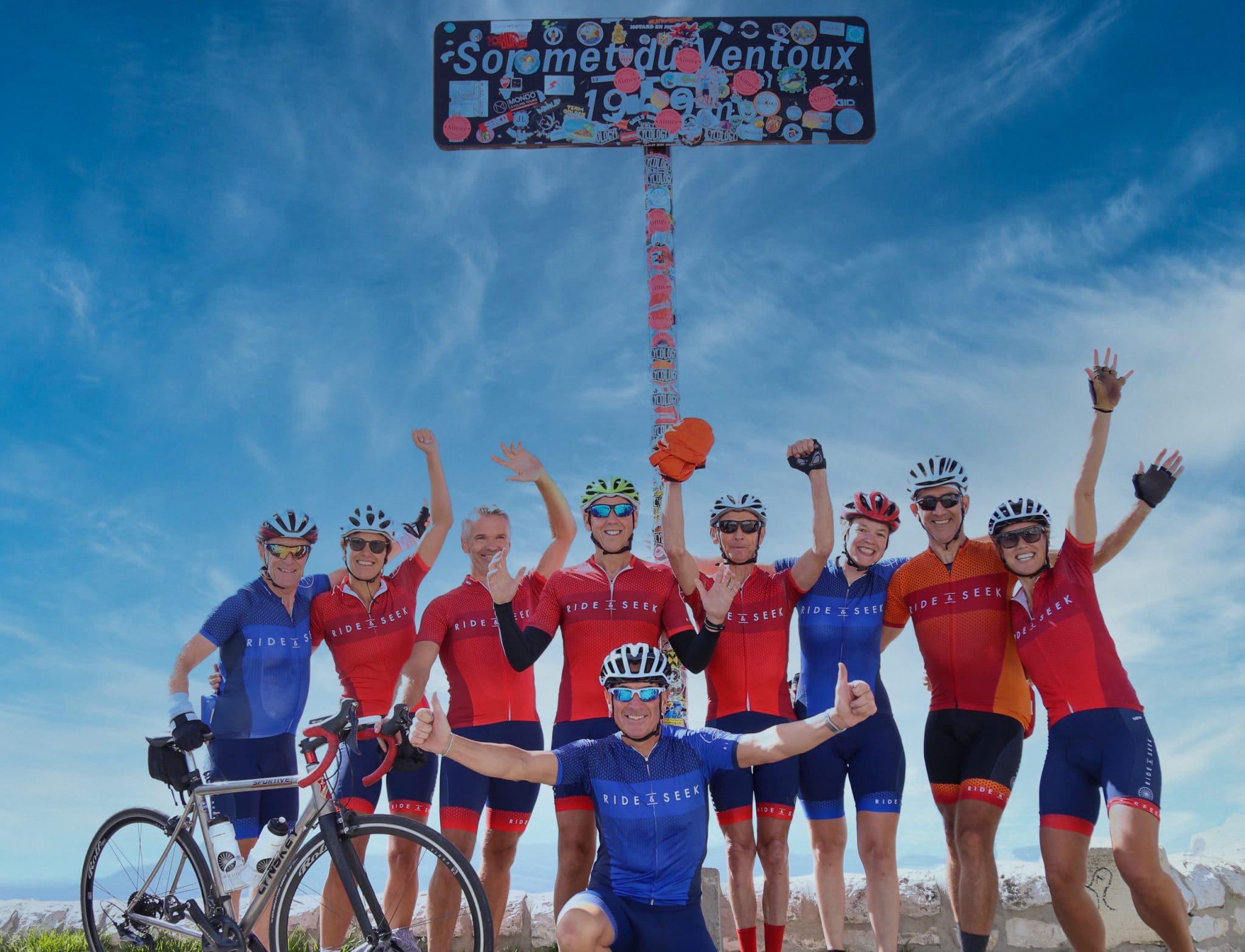 A group photo of cyclists at the summit of Mont Ventoux with arms raised in the air.