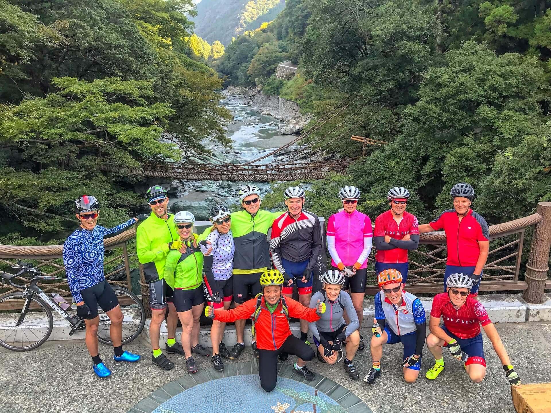 A group of Ride and Seek cyclists posing in a group photo on a bridge in Japan. nine riders are standing at the back and 4 are kneeling in front. There is a creek flowing under the bridge and a bike behind the group.