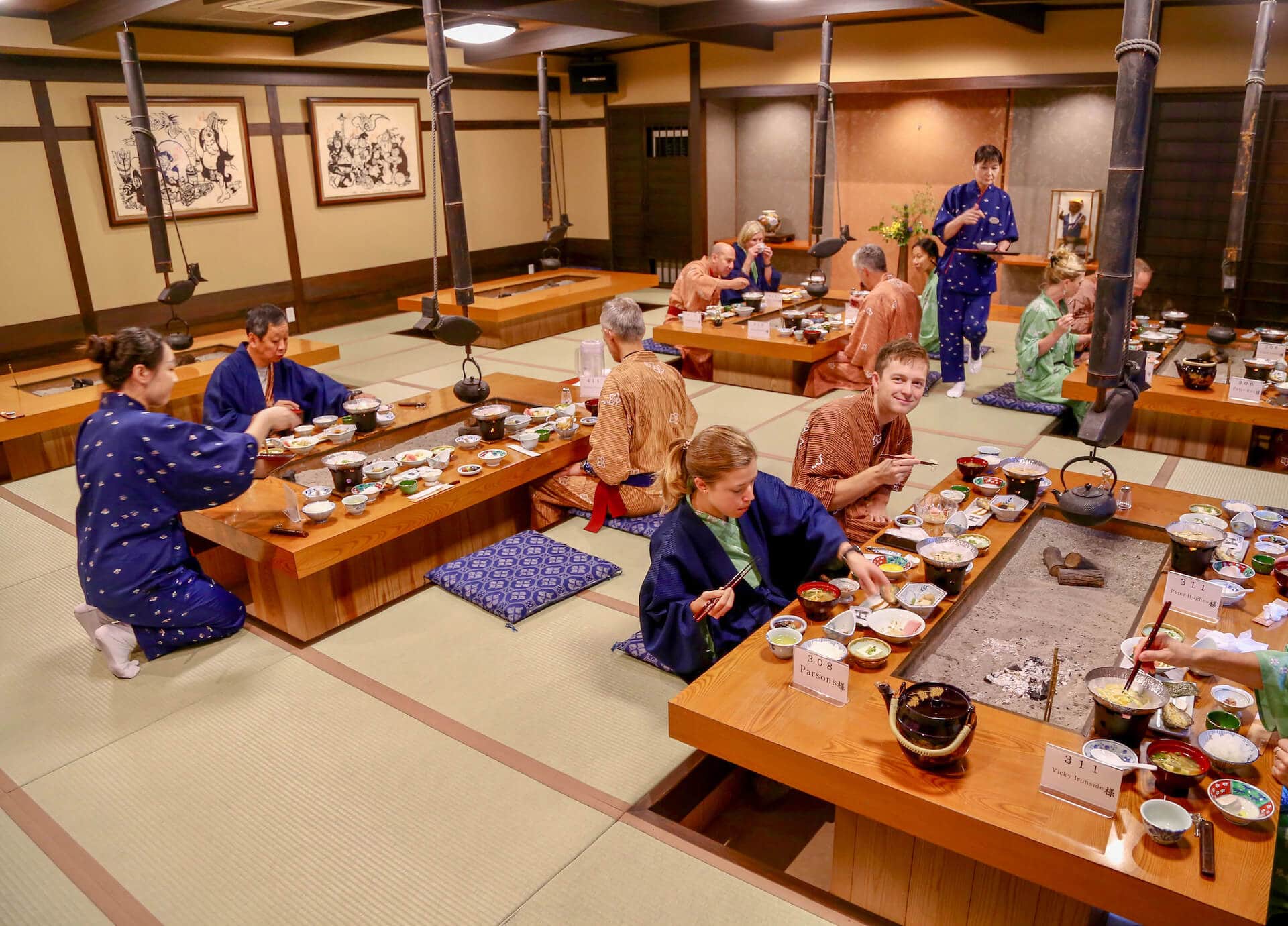 Ride and Seek guests seated on the floor around four separate tables in a Japanese eating house in traditional Japanese dress. The tables where the guests are seated are laid with many bowls of various Japanese dishes.