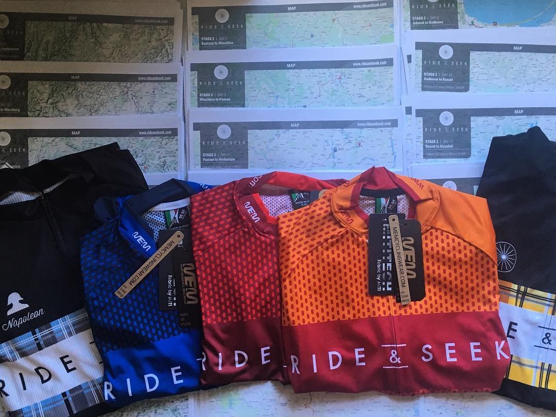 Photo of a variety of Ride and Seek jerseys is orange, red, blue and yellow tartan and blue tartan lying on some printed maps that show the Ride and Seek cycling tours routes.