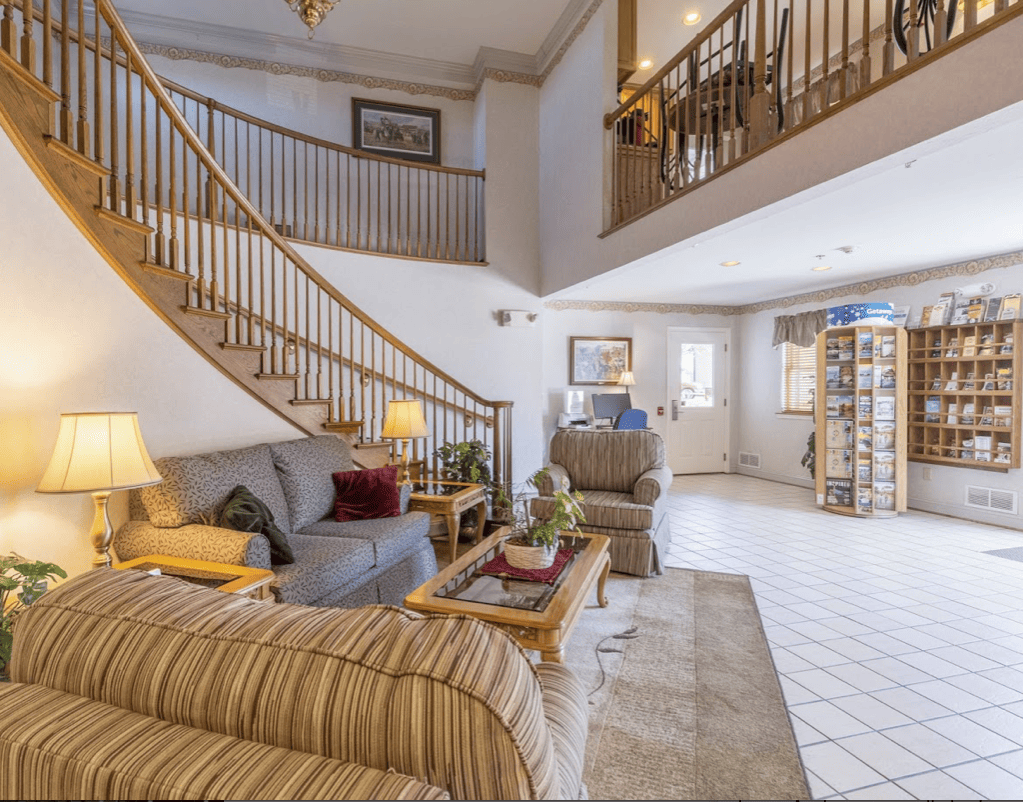 Inside the lobby of a hotel. Stylish lounge furniture is arranged in the centre of the lobby with a sweeping staircase rising up to an open upper floor level with balustrades along the edge of the upper floor..