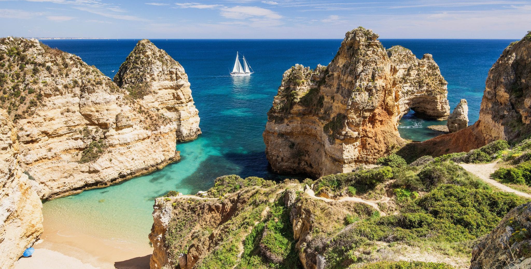 A tiny seaside beach is nestled among tall outcrops and deep blue ocean. A white yacht under sail is on the water just offshore.