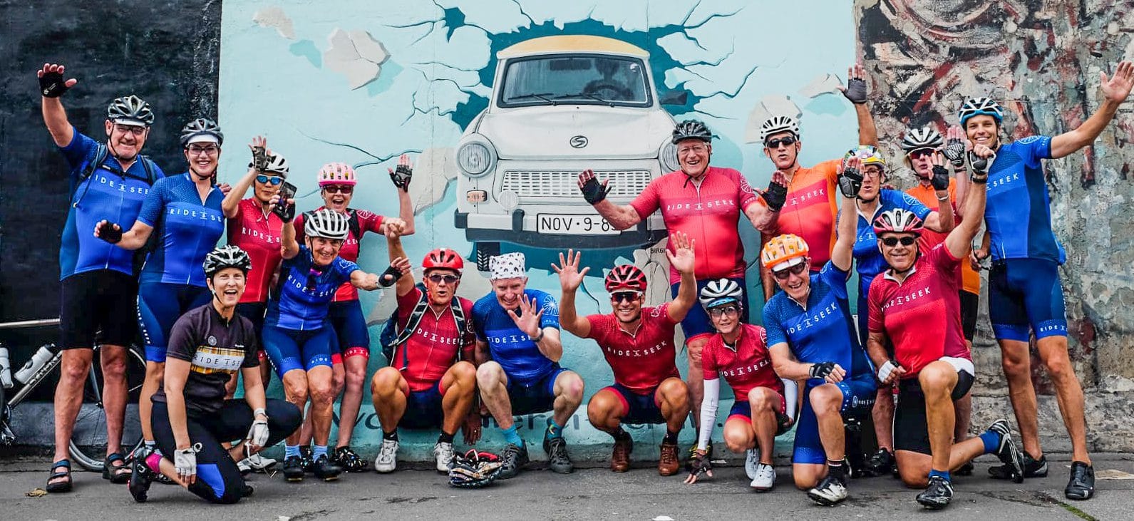 A group of cyclists dressed in various Ride and Seek cycling kits pose in front of a Berlin wall mural of a car appearing to break through the wall.