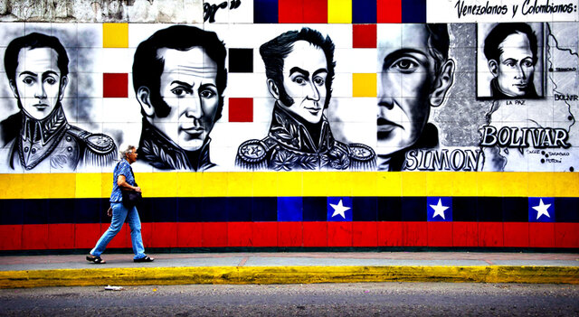 A person walks along a street with a mural depicting a man in several different poses along the wall. The mural has the words Simon and Bolivar written in capital letters under one of the faces. Under the faces are three stripes. The top one is yellow, the middle one is black with three white stars in a blue box. The bottom stripe is red. Red, yellow and black squares are dotted between the faces on the wall.