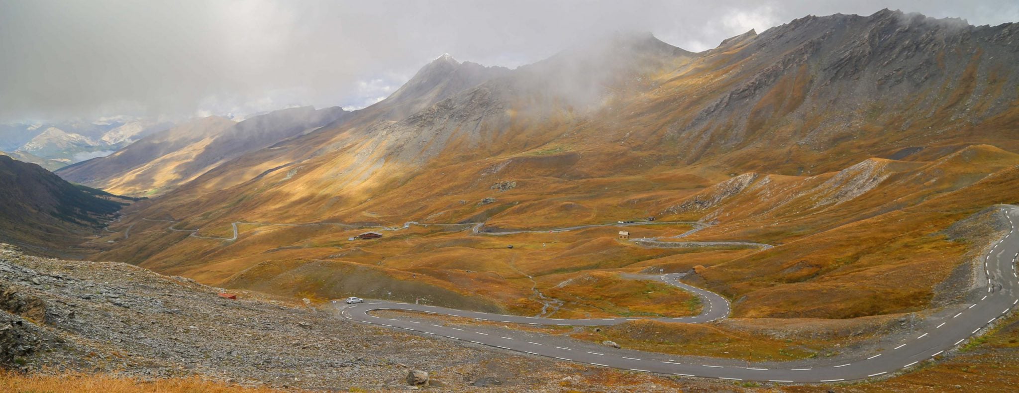 A high mountain road winds down a dry valley with tall mountain ridges. Many switchbacks are visible.
