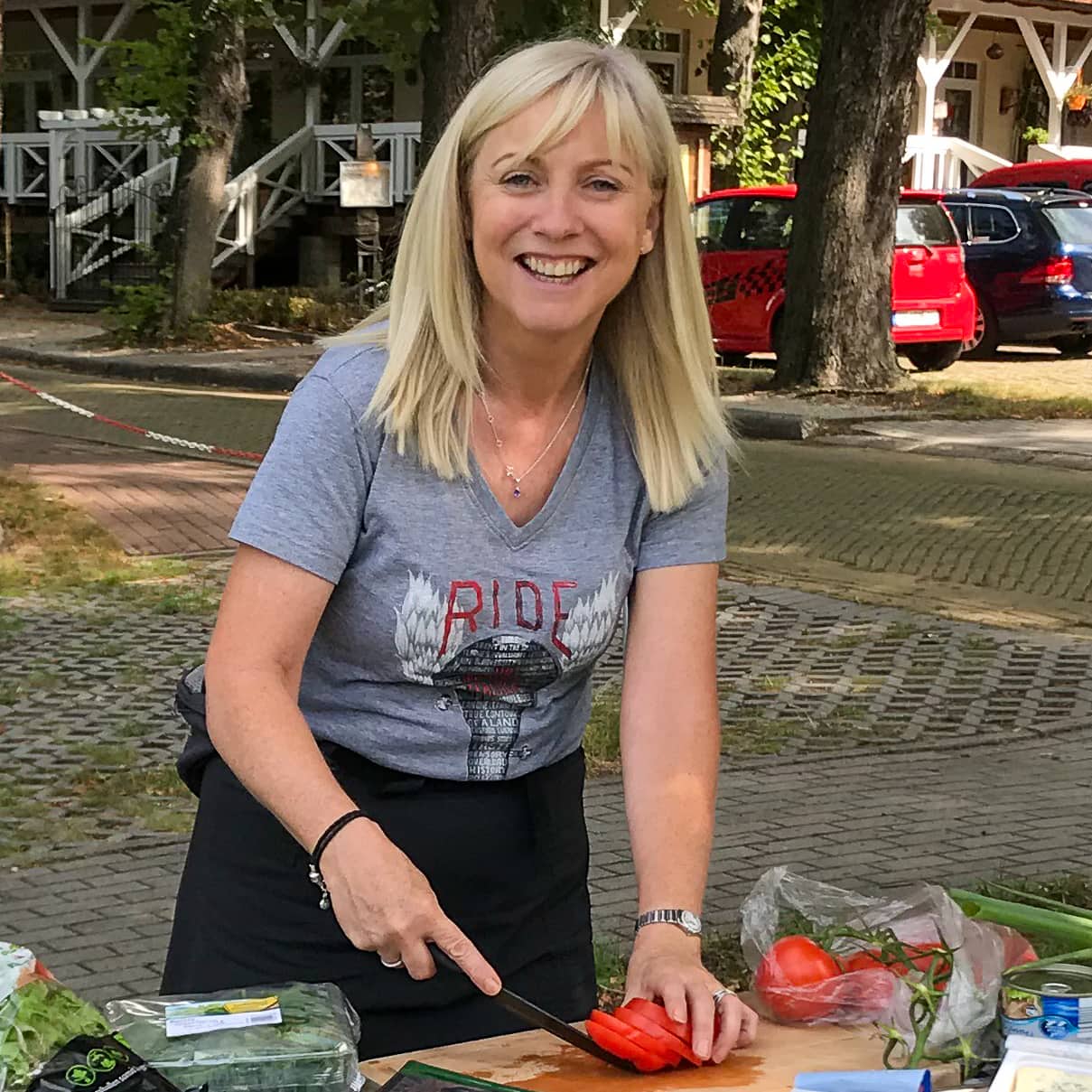 A smiling woman with blond hair wearing a t-shirt with ride and seek cycling tours printed on the front. The woman is holding a knife and slicing a tomato. There is other fresh produce on the table. The scene is set outside in a street with cars in a car park across the road.