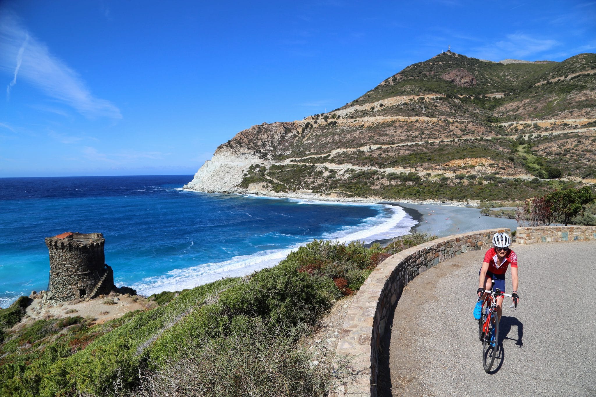 A cyclist rides out of a corner of a road lined with a rock wall, along the seaside. Below is a grey beach, an old round tower and a tall hill in the background.