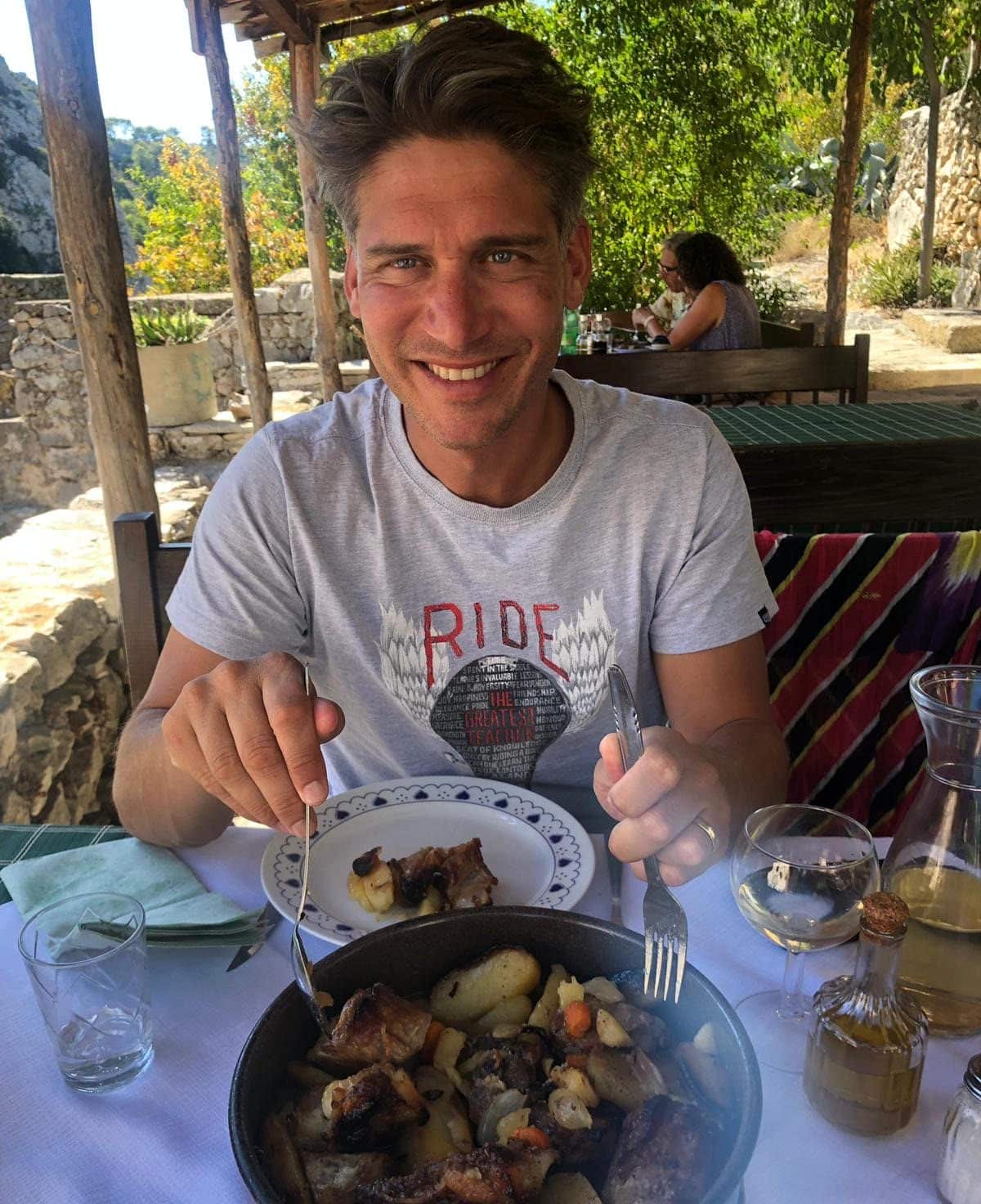 A smiling person sits at a table about to serve themselves from a round dish in an alfresco, Mediterranean setting.