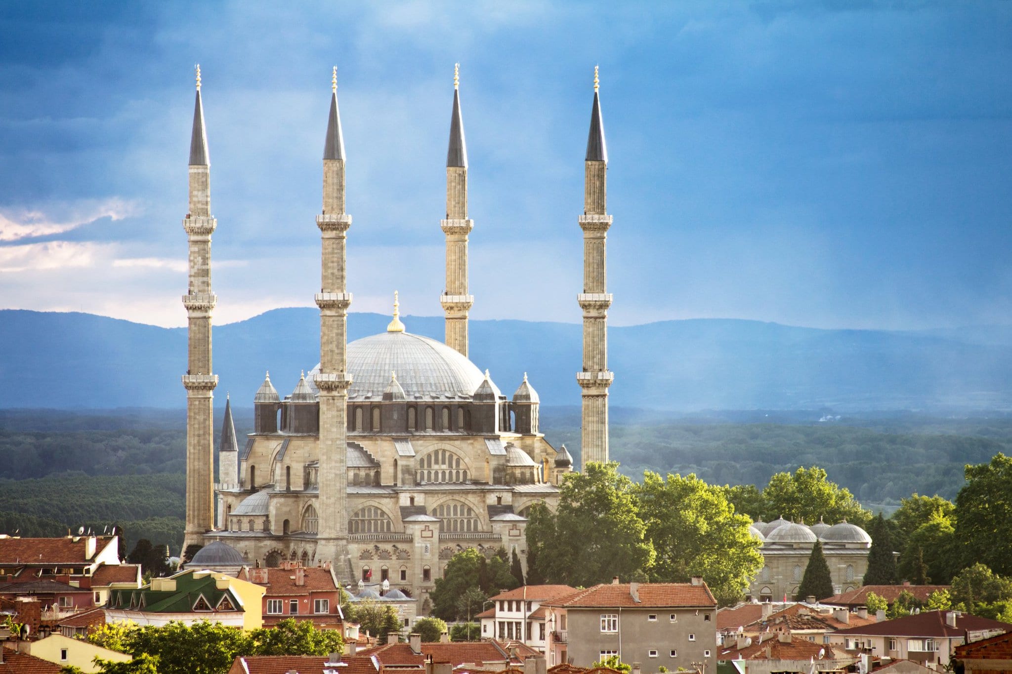 A large white mosque with four minarets stands tall, sitting within an old twon with red teracotta roofed houses. A mountain range stretches across the background.