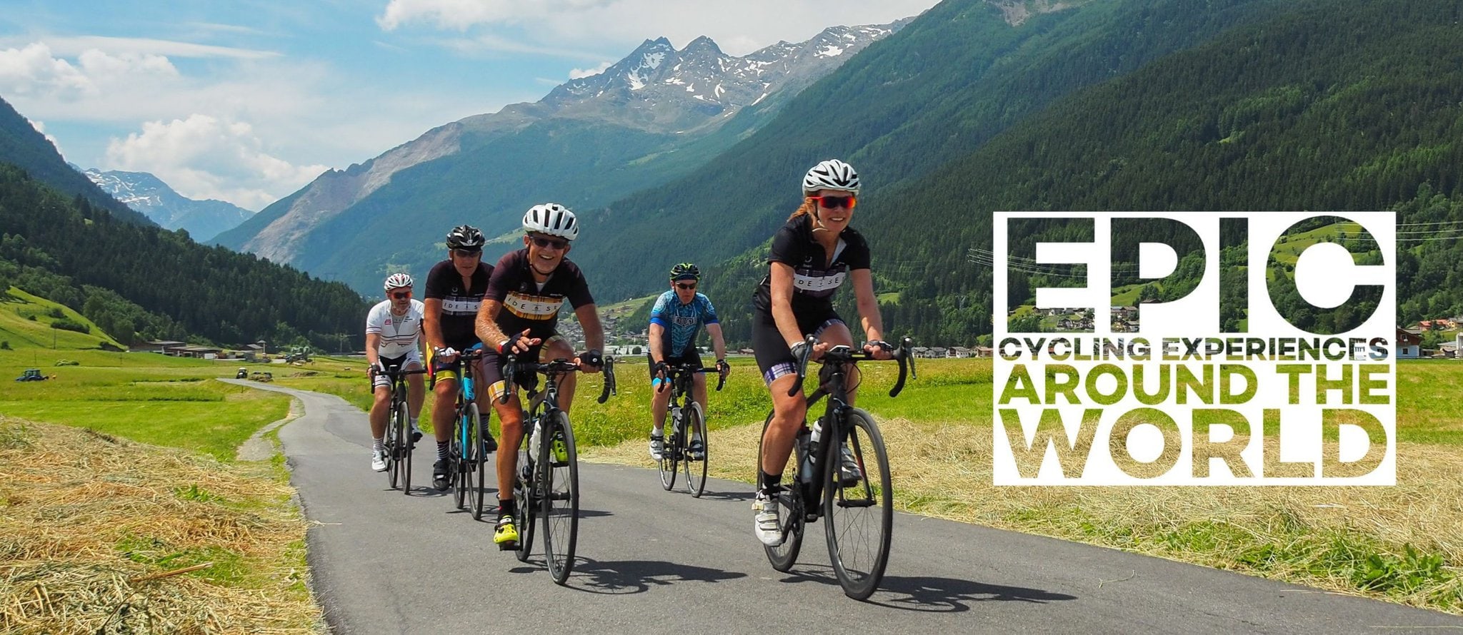 A group of Ride and Seek cyclists riding along a quiet road in a valley with snow covered peaks in the background.