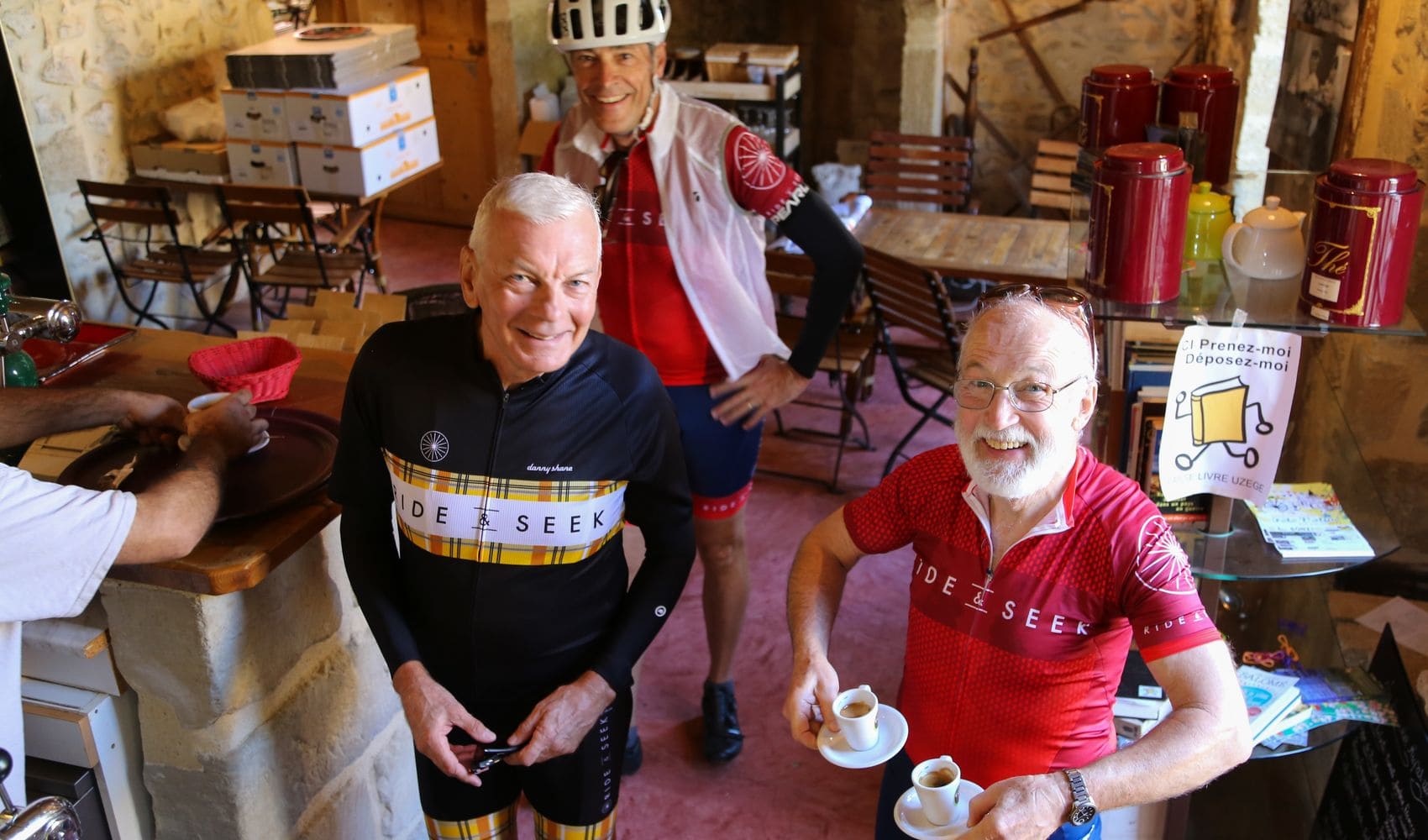 Three cyclists wearing various Ride and Seek kits taken from a higher vantage point stand in a coffee shop. One is holding two small espresso cups of coffee. The bar attendant can be partially seen preparing another cup on the counter to the side.
