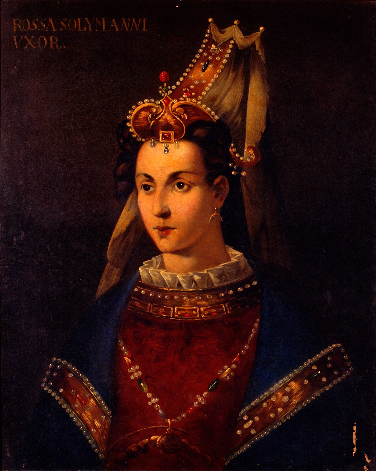 A painted portrait. The subject wears an elaborate headdress adorned with jewels and pearls. The clothing is red with a ruffled white collar, adorned with more pearls. Over the clothing is a long, jewelled necklace and a blue adorned cloak.