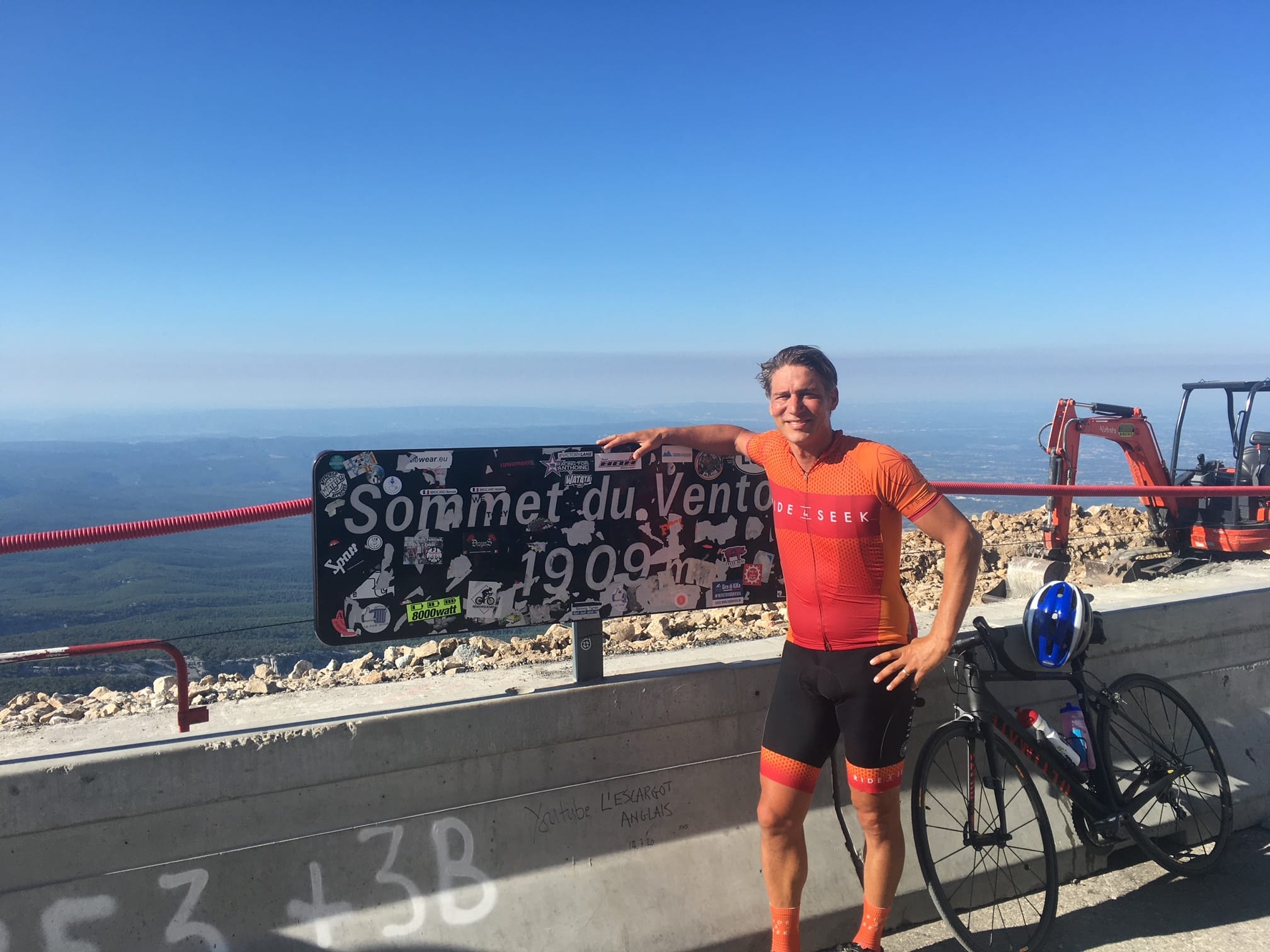 Dylan poses with his bike, resting against a summit sign. A vast expanse of water stretches off into the distance.
