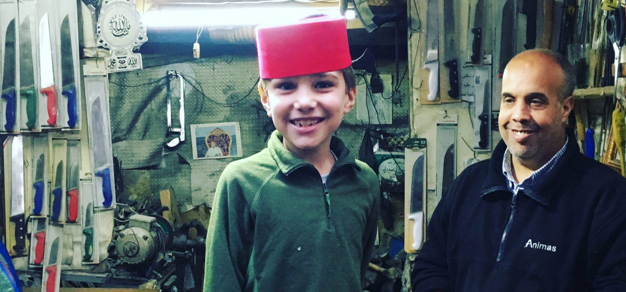 A boy stands smiling, wearing a red round hat. A man stands to the right of the boy, smiling at him. F