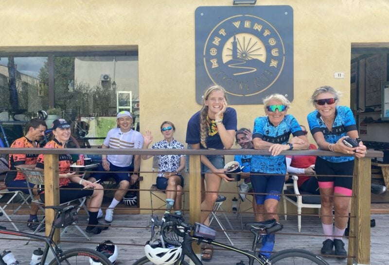 A group of three people dressed in cycling gear stand leaning on a balcony rail. another group sit on seating behind them.