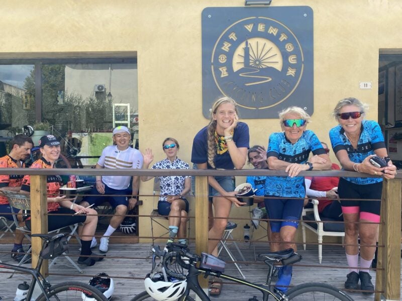 A group of three people dressed in cycling gear stand leaning on a balcony rail. another group sit on seating behind them.