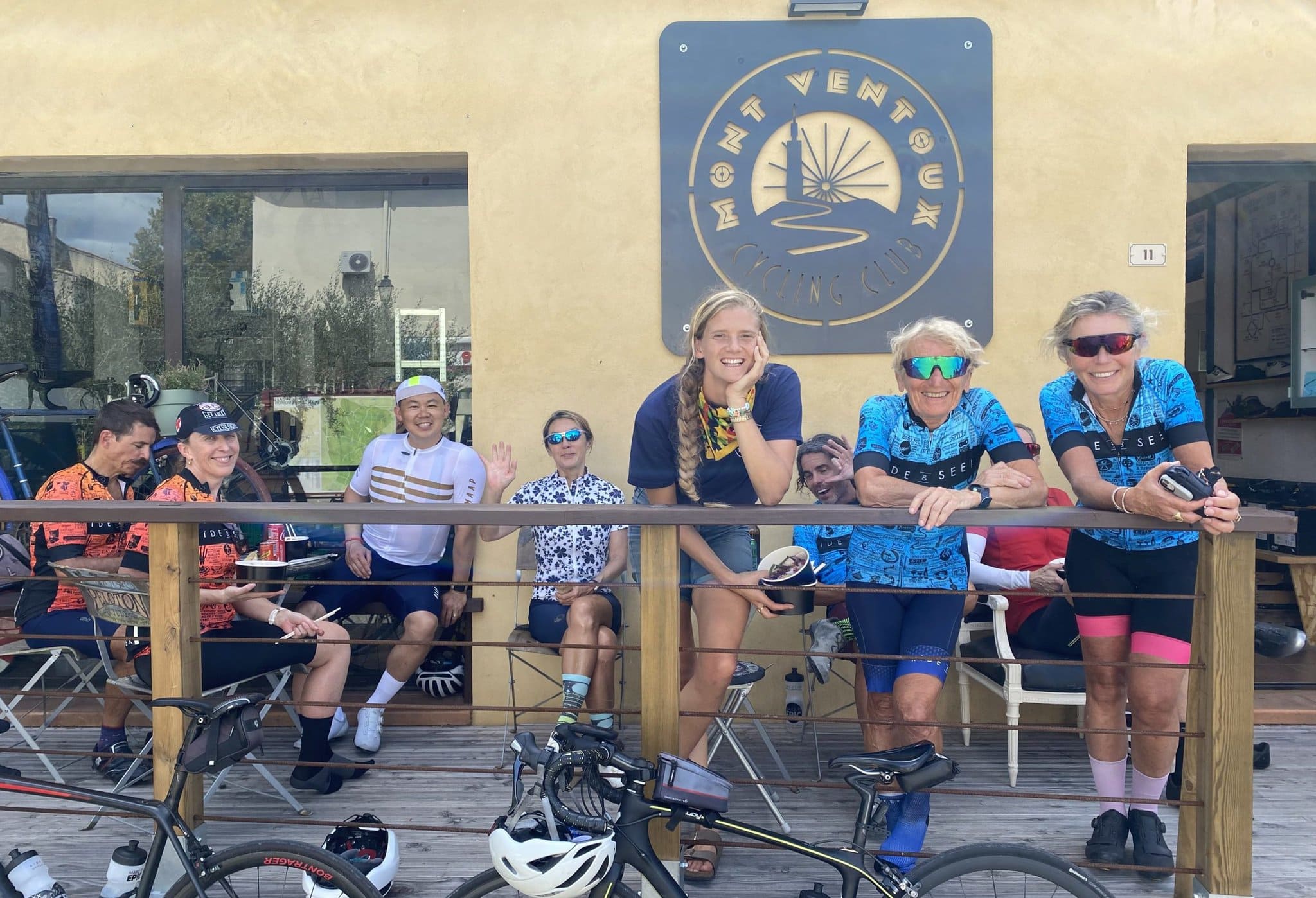 A group of cyclists on a patio. A sign is hanging on the yellow wall behind them. The lettering reads Mont Ventoux Cycling Club. Three cyclists are standing in the front, leaning on the railing. Six are seated behind.