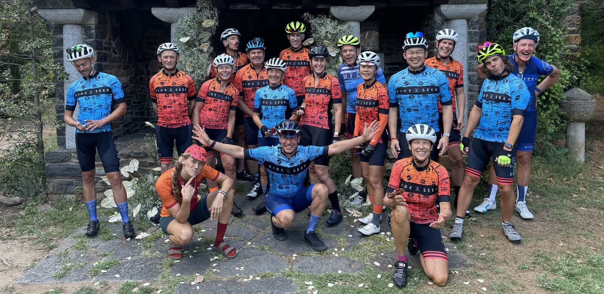 A photo of a group of riders in Ride and Seek cycling tours kit wearing a riding helmet in various poses. Three cyclists are kneeling, one of whom has their arms spread wide.