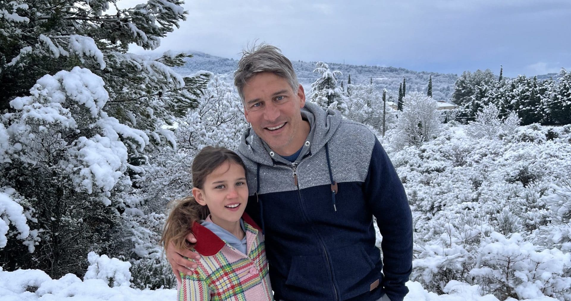 A man and a young girl pose together, set in a snowy landscape. Behind them are snow-covered trees and bushes.