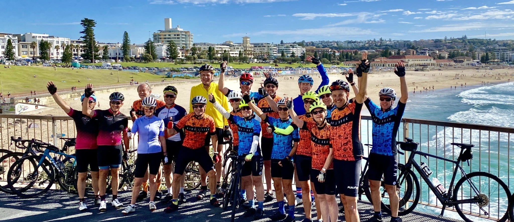 A group of Ride and Seek cycling tours guests pose with their arms raised in front of a sandy beach on a sunny day. The riders' bikes lean against the safety fence behind the group with city buildings in the background. .