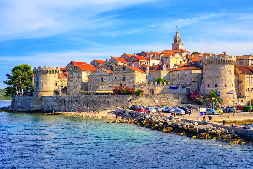 Home of Marco Polo, an old town built on the water's edge, with a round tower at each of the two visible corners. People and cars can be seen behind and in front of the protective wall built out into the water. 