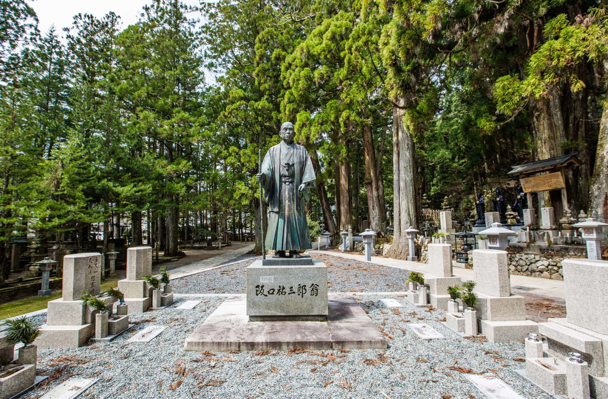 A stone statue sits in a clearing among headstones in a park with vibrant green trees in the background.