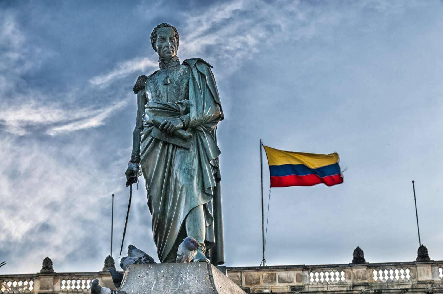 A statue of a man in military attire with a long cape draped over his shoulders and wrapped around his waist. A country flag with yellow, blue and red horizontal stripes flies on a pole from the top of the building behind the statue.