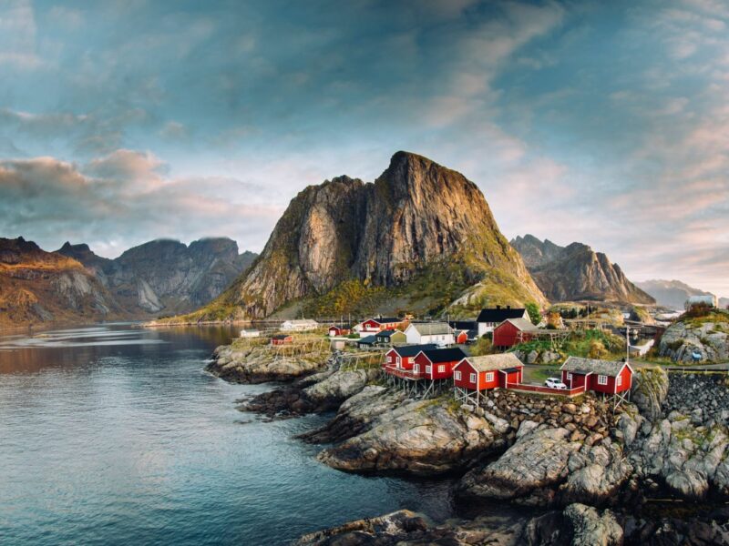 A small coastal village with red cottages that are built on stilts over the rocky edge of the water. A tall rocky outcrop partially covered by grass in the background.