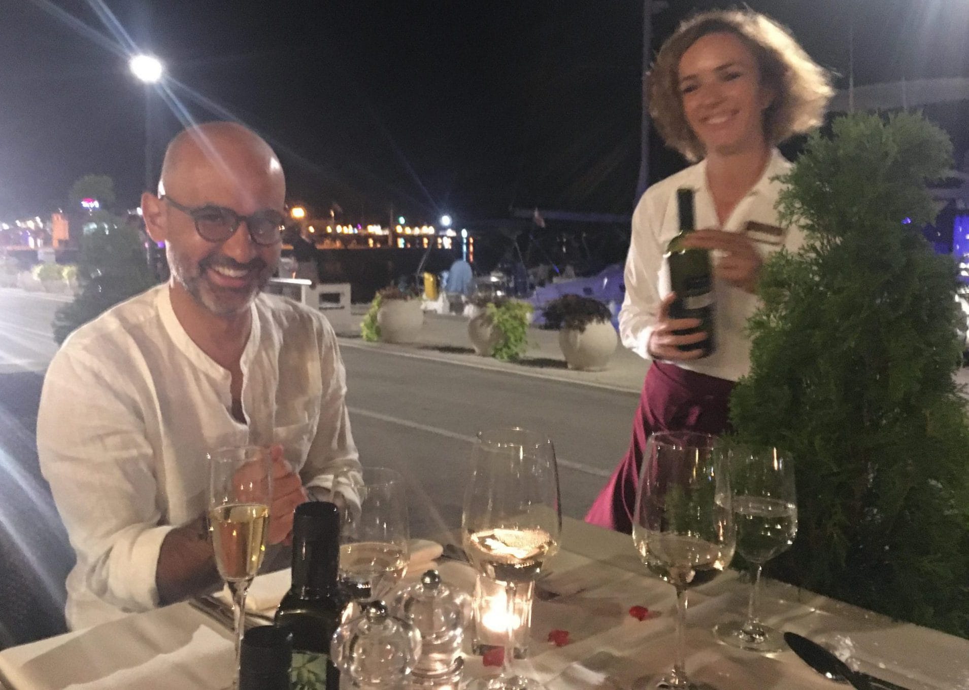A wait staff holding a bottle of wine stands beside a seated guest at a table set with glasses and a lit candle. A camera is sitting on the table.