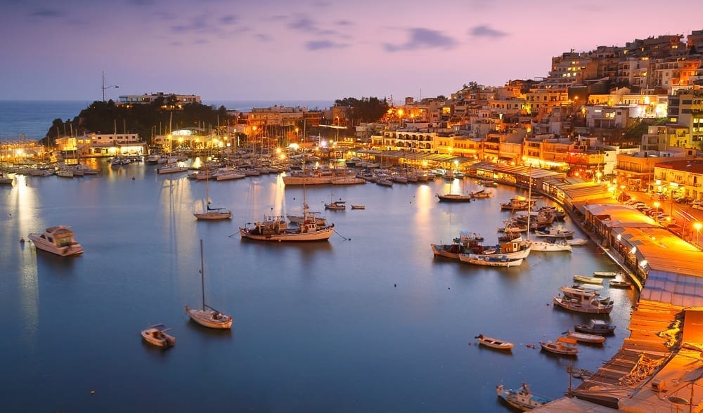 A town at night is built around a small bay. The streets are lit in golden light, and boats are moored in the water. The sky has a purple and pink glow.
