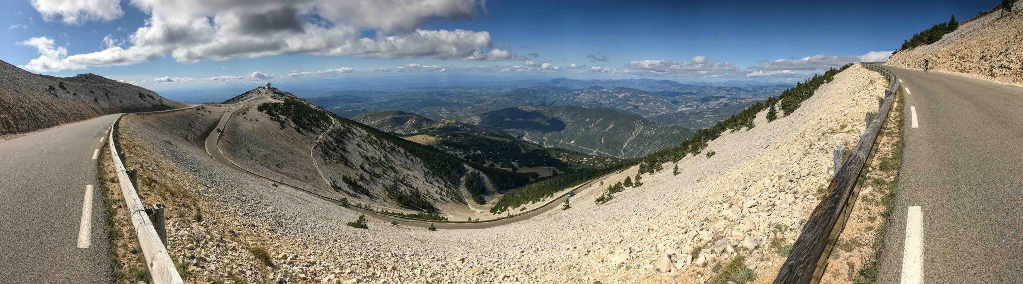 Panoramic view of a mountain road taken from the bend of a switchback. The valley below is backed by a blue, cloudy sky.