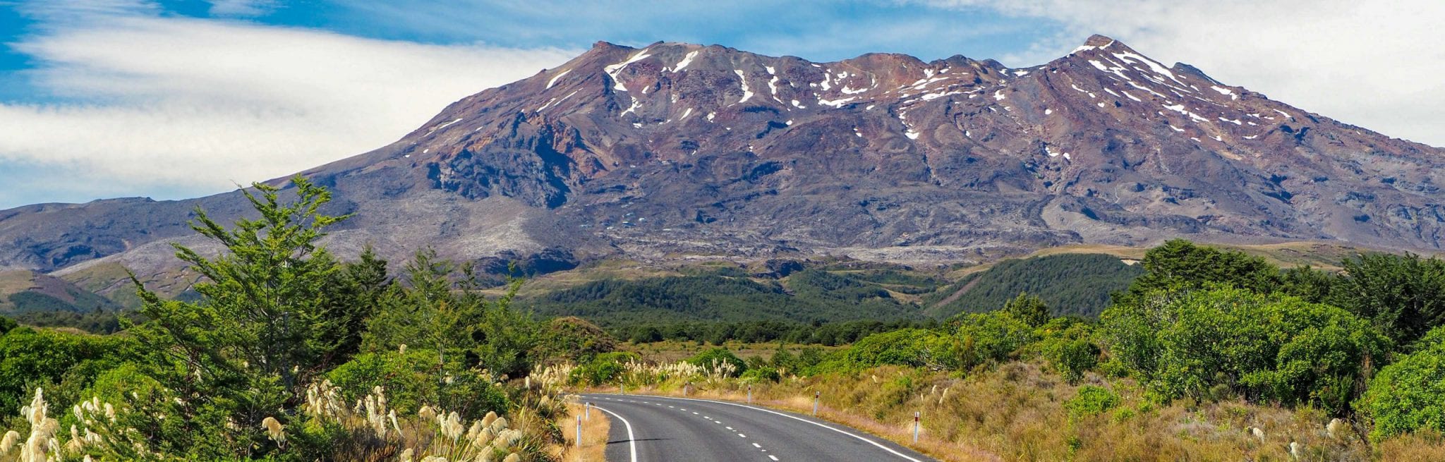 A road leads towards a tall, snow-covered mountain in the background. A blue, cloudy sky is behind the mountain.