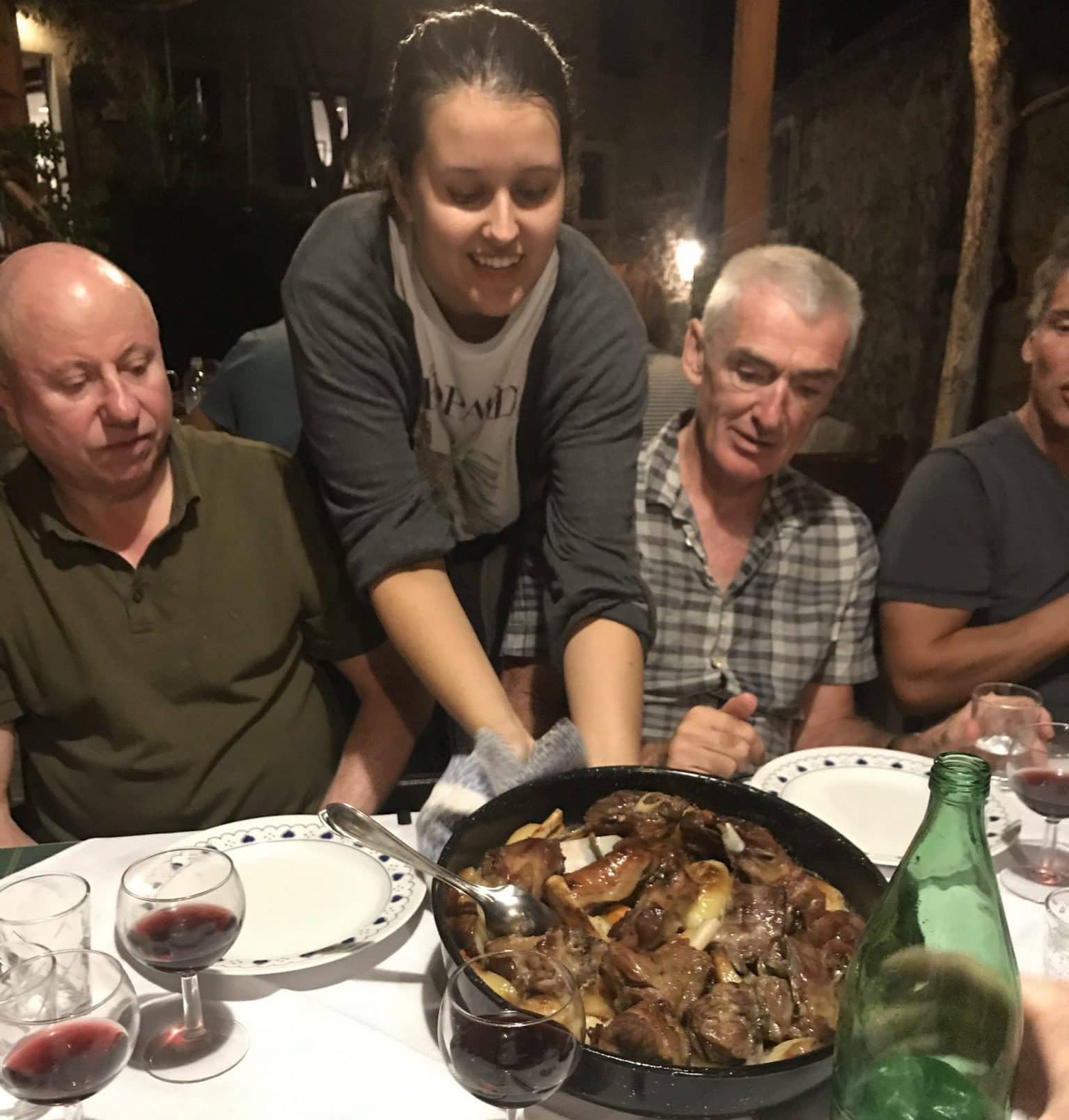 A wait staff member leans down between two seated guests who are watching on. On the table is a large round dish of food ready to be eaten. Glasses of wine and plates are set on the table.