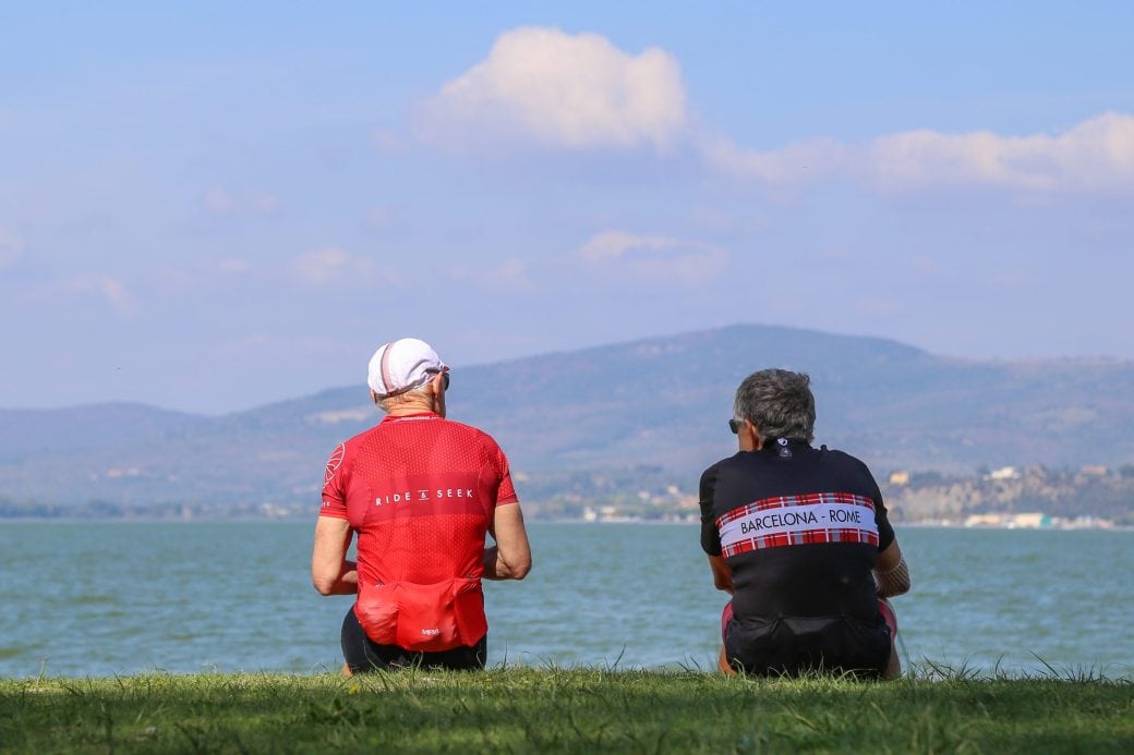 Two people wearing Ride and Seek cycling tours jerseys sitting on green grass, face away from the camera and look out on a lake with a town and mountains in the background.