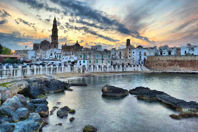 An old town built on the water's edge. An old church with a tall steeple is nestled behind houses. The sunlight is fading with sunset colours in the sky and streaky cluds.