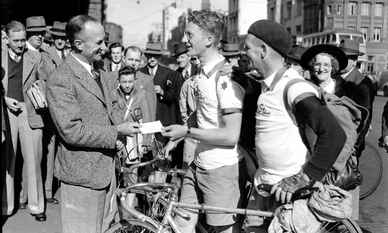 A historical sepia photo of a man handing a ticket to a cyclist with another cyclist standing behind, holding a vintage bike. There is a crowd of people in the city street, watching in the background.