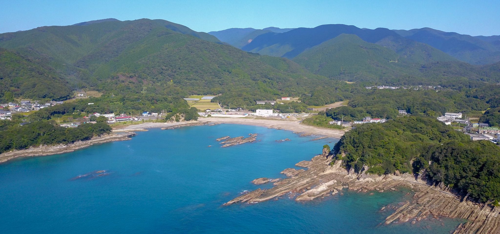 A craggy shore in front of a wide beach taken from a high vantage point. There are hills in the background set against a blue sky.