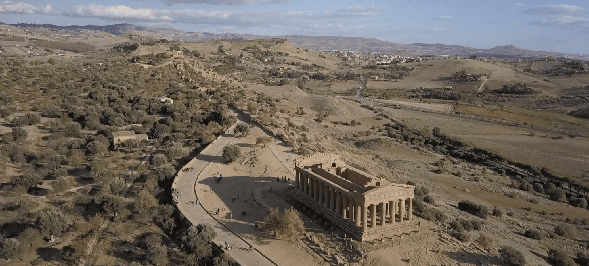 An aerial photo of an ancient, roofless building, set on a dry landscape with hills in the background. A road rings around the side of the building.