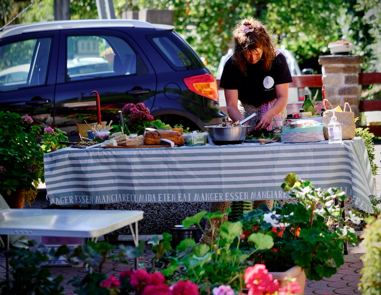 A Ride and Seek bike tours guide prepares picnic food on a table. The table is laid with picnic food on a blue and white striped tablecloth. In the background there is a small blue car. There are bright flowers and greenery in the foreground.