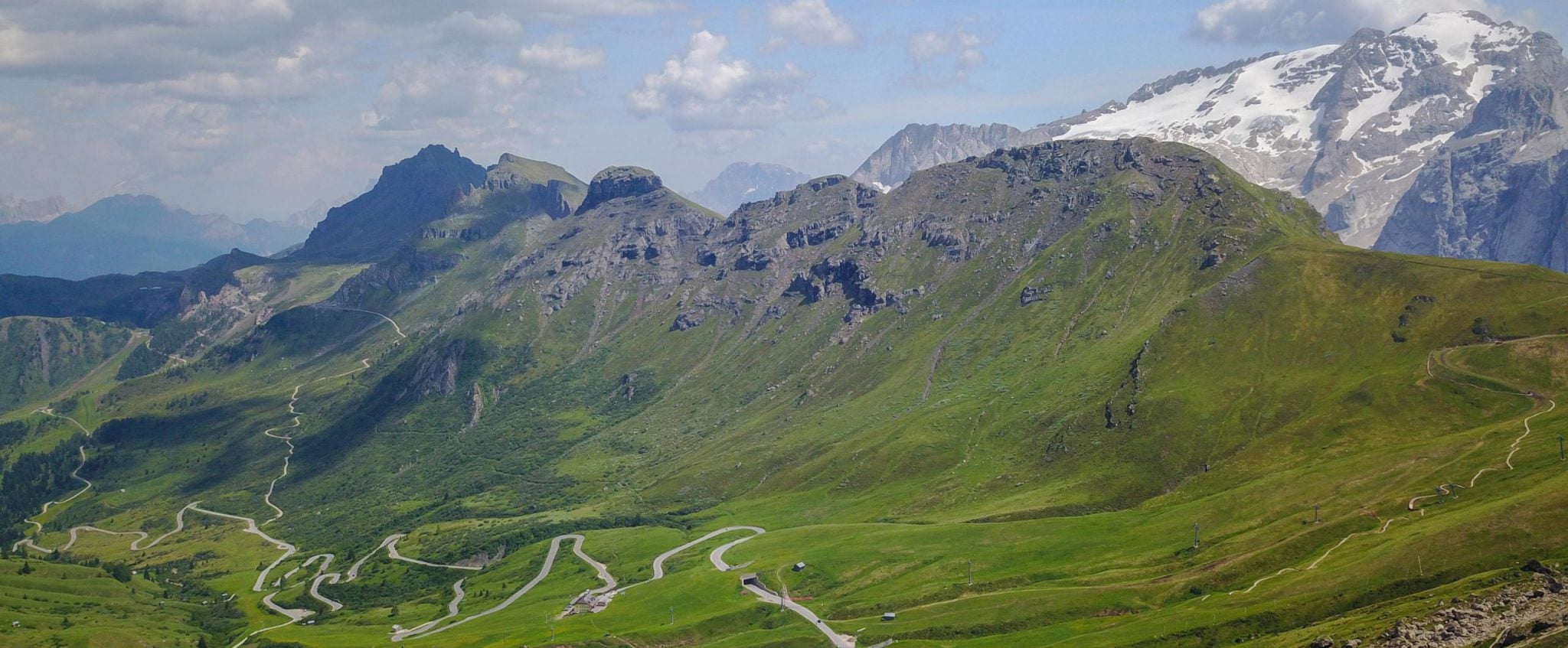 A road winds down through a valley with snow covered alps in the background.