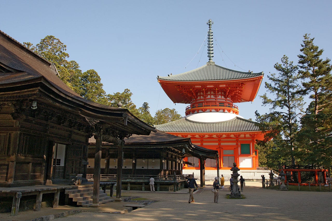 A photo of a red temple with one ornate roof sitting atop a second and a tower protruding from the top roof. Brown buildings in the foreground.